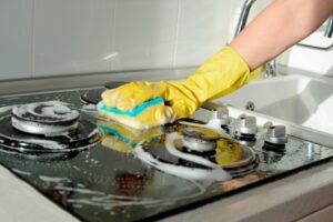 A hand in a yellow rubber glove washes a gas stove on a sunny day. Kitchen cleaning